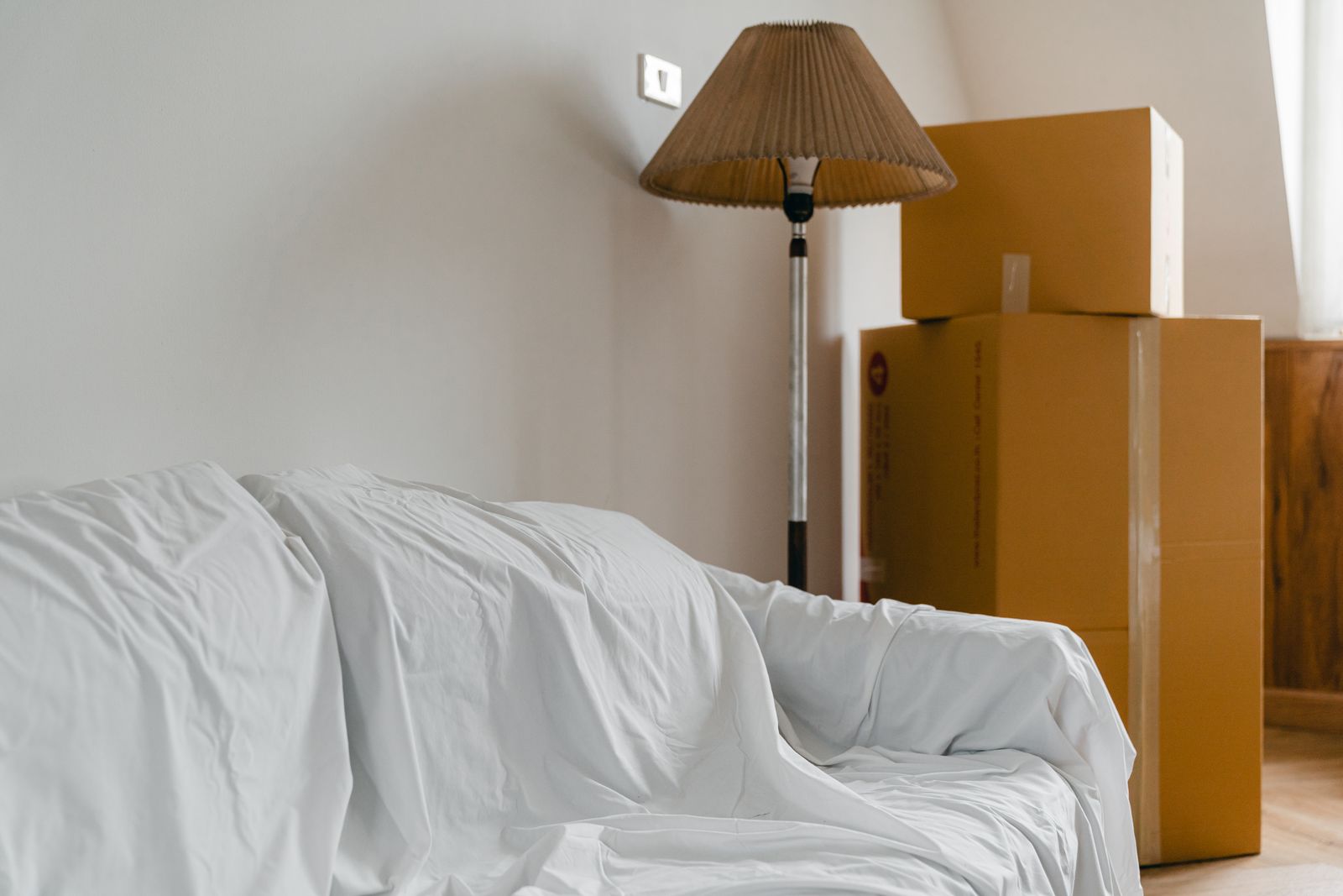 Packed and labelled moving boxes beside a sheet-covered sofa in a bright lounge room